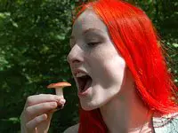 Woman with vivid red hair moving a red mushroom towards her open mouth.